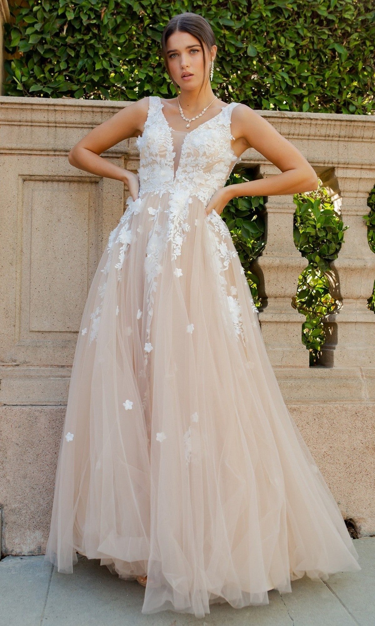 A woman stands outdoors in front of a stone railing, wearing the Cinderella Couture 8040J long formal dress in champagne, featuring a sleeveless design, elegant 3D floral lace appliques, and a flowing tulle floor-length skirt.